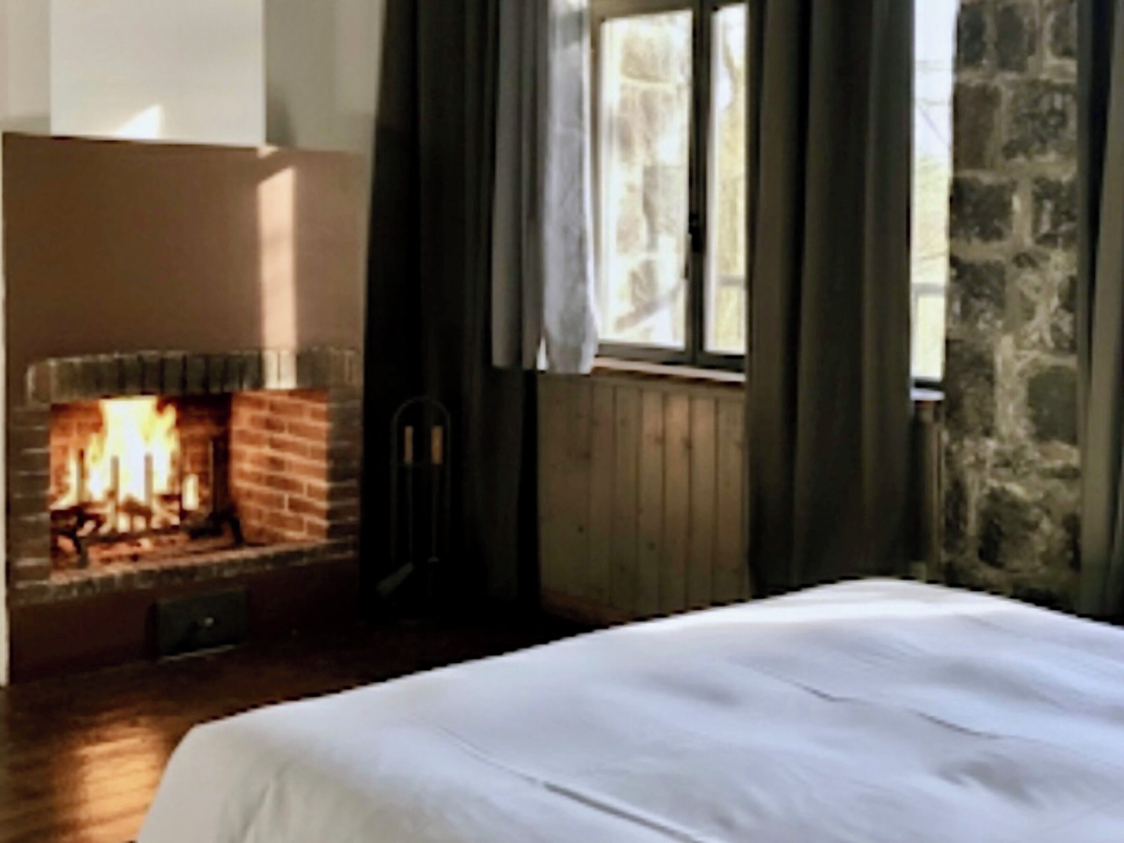 Een kamer met een schouw van baksteen, houten vloeren, een bed met wit beddengoed en een raam met gordijnen.