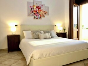 A neatly made bed with white linens, two bedside tables with lamps, and a floral painting above the bed. - B&B Villa Loriana (Milo)