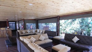Un espace extérieur aménagé, abrité et doté de canapés et de coussins en osier, d’un plancher en bois et de grandes fenêtres offrant une vue sur l’extérieur.