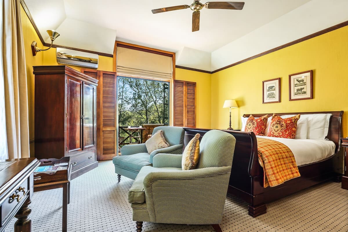 A bedroom with a bed, a green armchair, a wooden dresser, and a window with a view of trees.