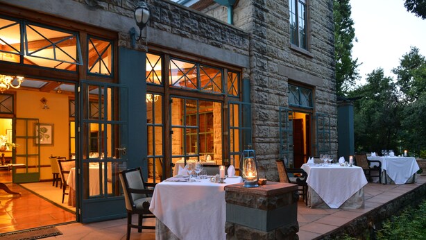An outdoor dining area with stone walls and large windows, tables set with white tablecloths and candles, and a lantern on a stone ledge. - De Hoek Country Hotel (Magaliesberg)