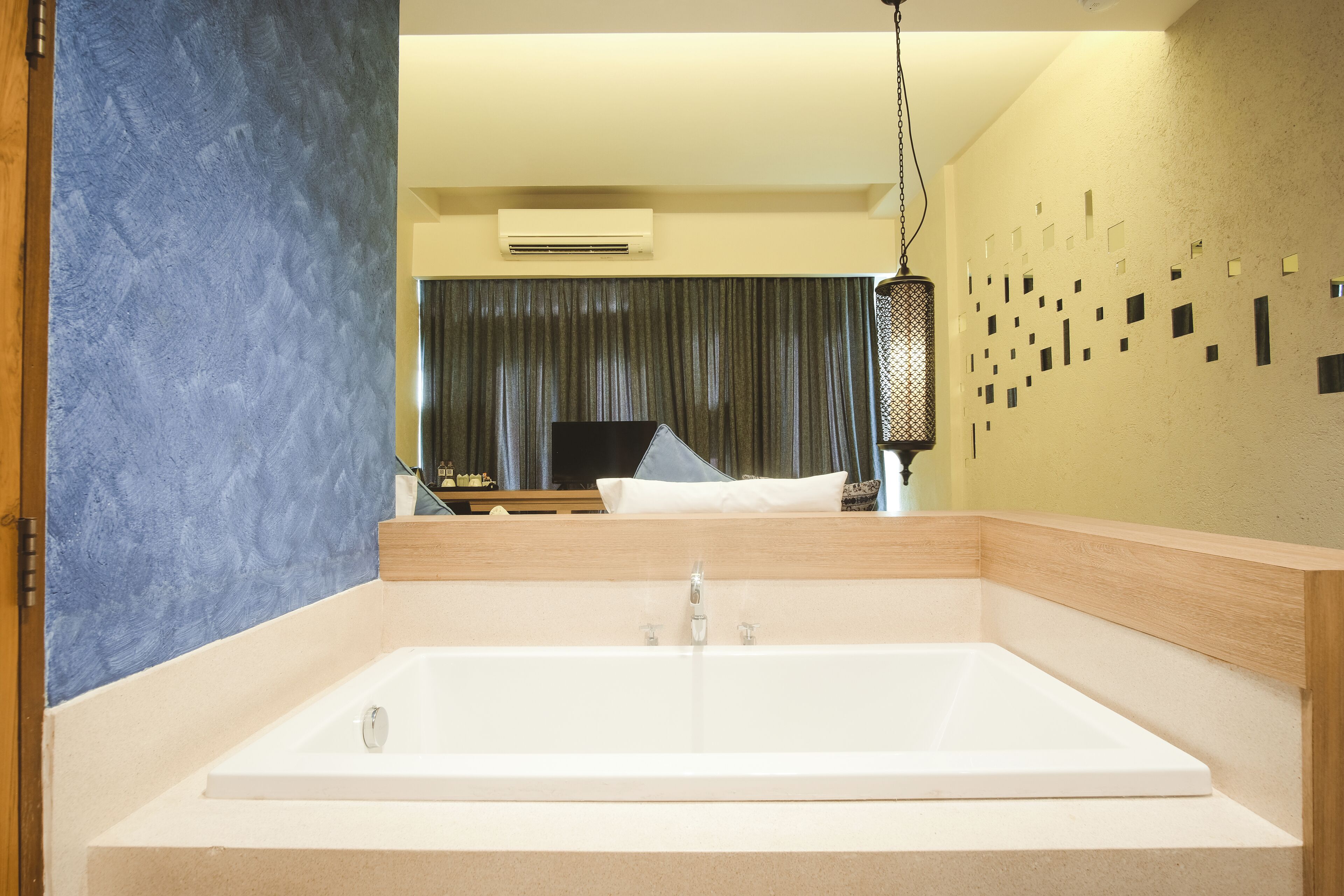 A modern bathroom with a large bathtub, a wooden vanity, and a mirror reflecting a living area with a television and curtains.