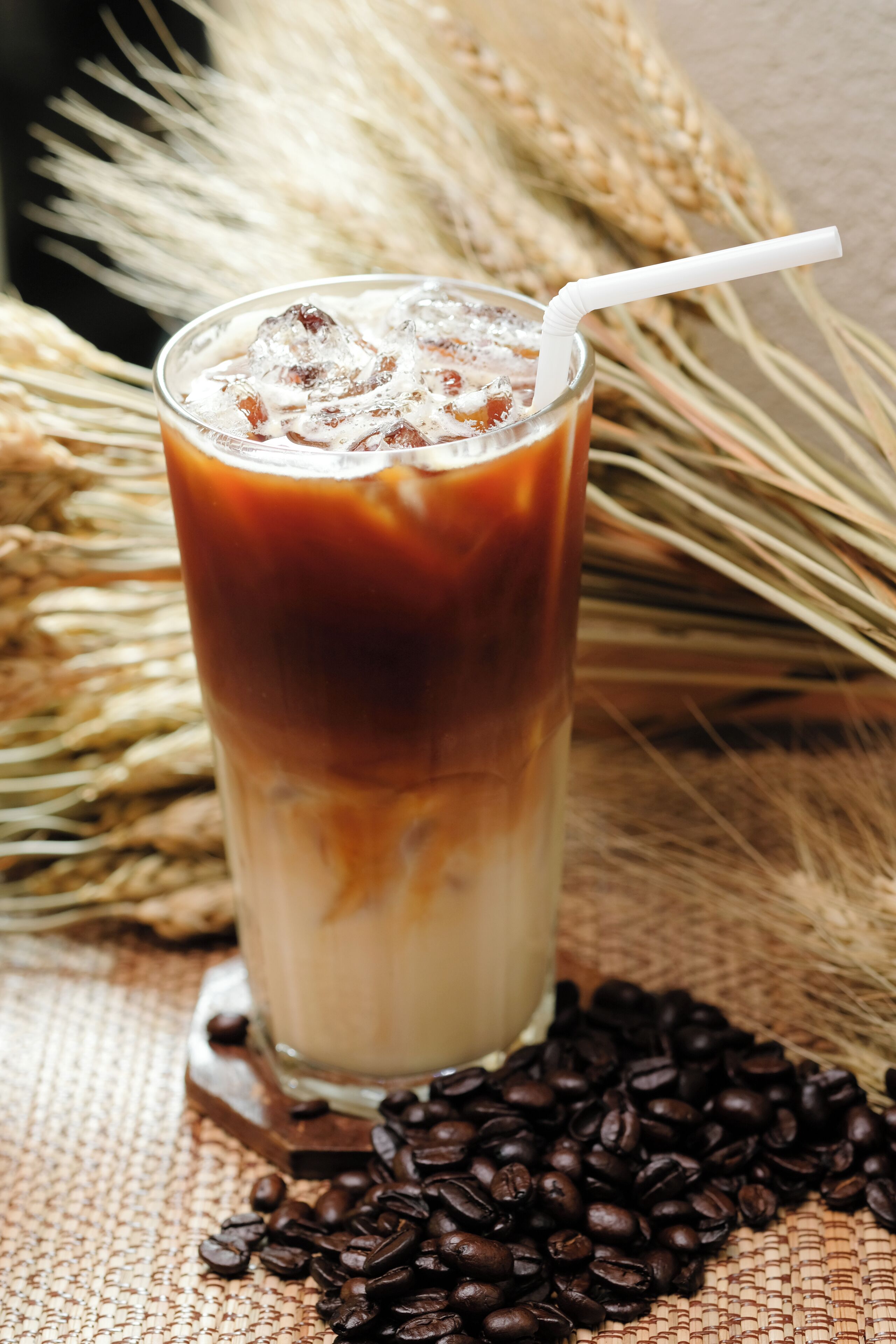 A tall glass of iced coffee with a straw, surrounded by coffee beans and wheat stalks.