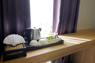 A tray with a fan, cups, a kettle, and a basket with toiletries on a wooden counter near a window with curtains.