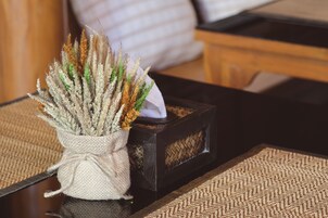Un jarrón rústico con plantas secas sobre una mesa de madera, junto a una caja negra y un mantel tejido.