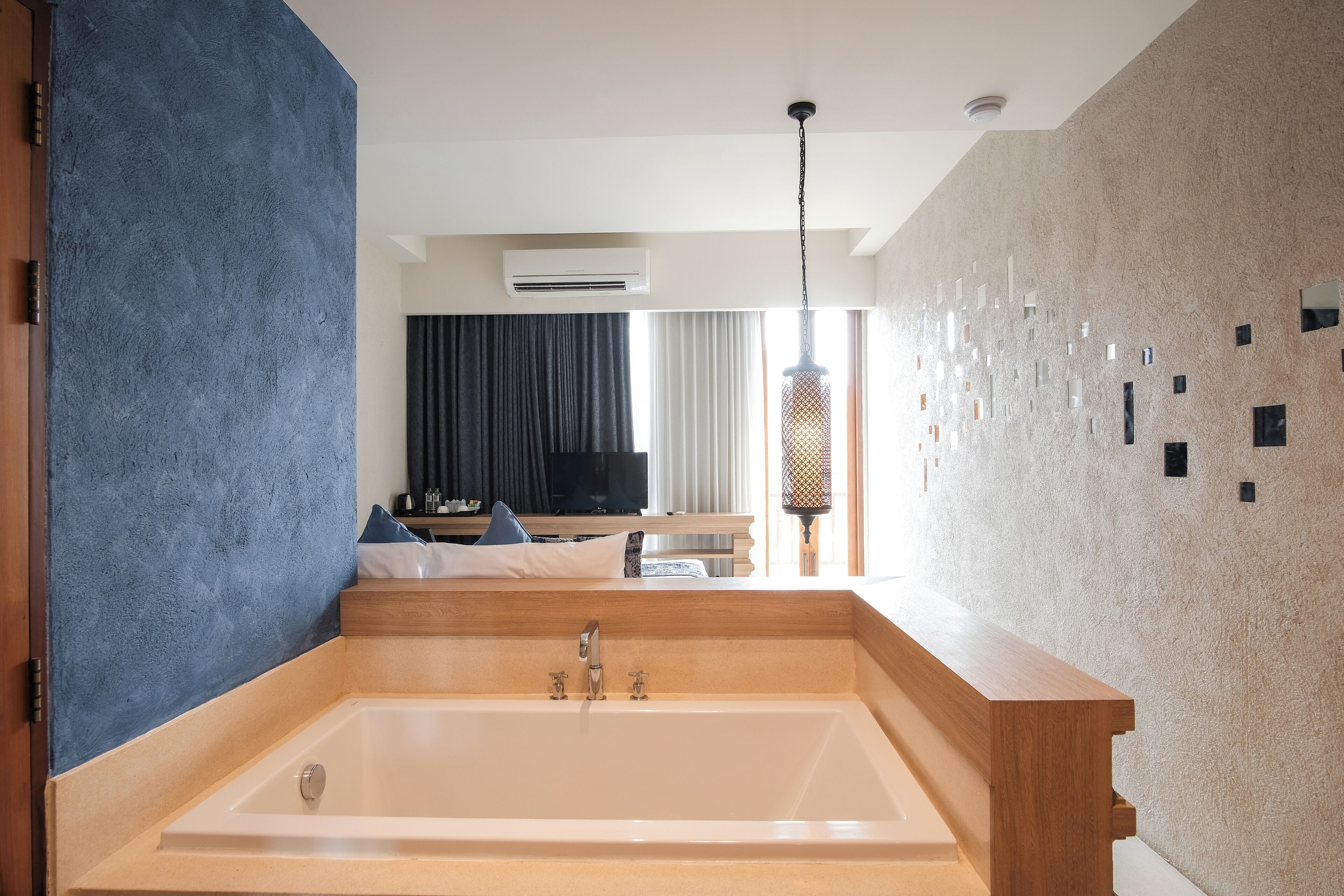 A modern bathroom with a large bathtub, a wooden bench, and a view of a living area with a television and seating.