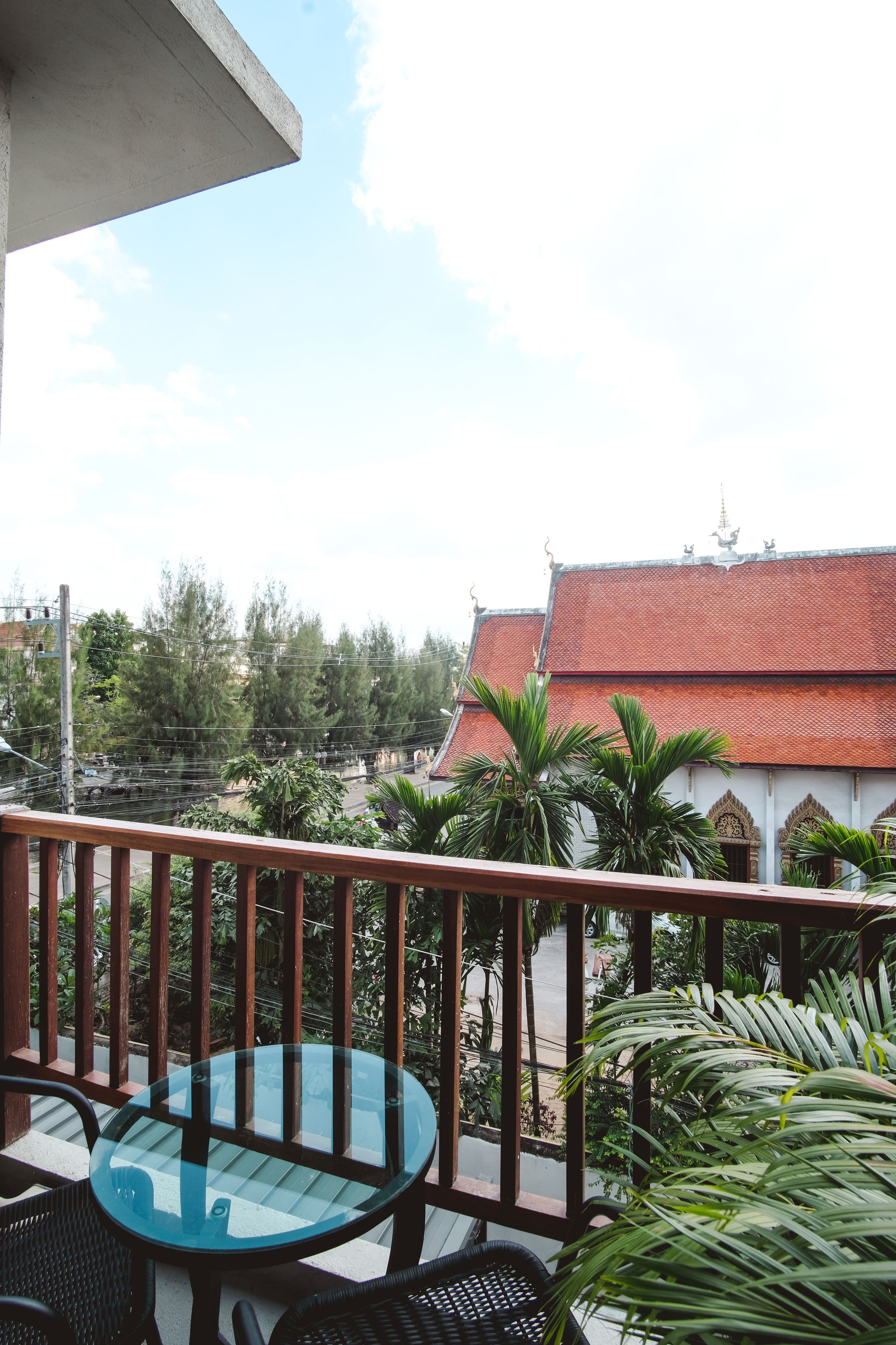 A balcony with a glass table and chairs, a red-roofed building in the background, and a view of the surrounding area.