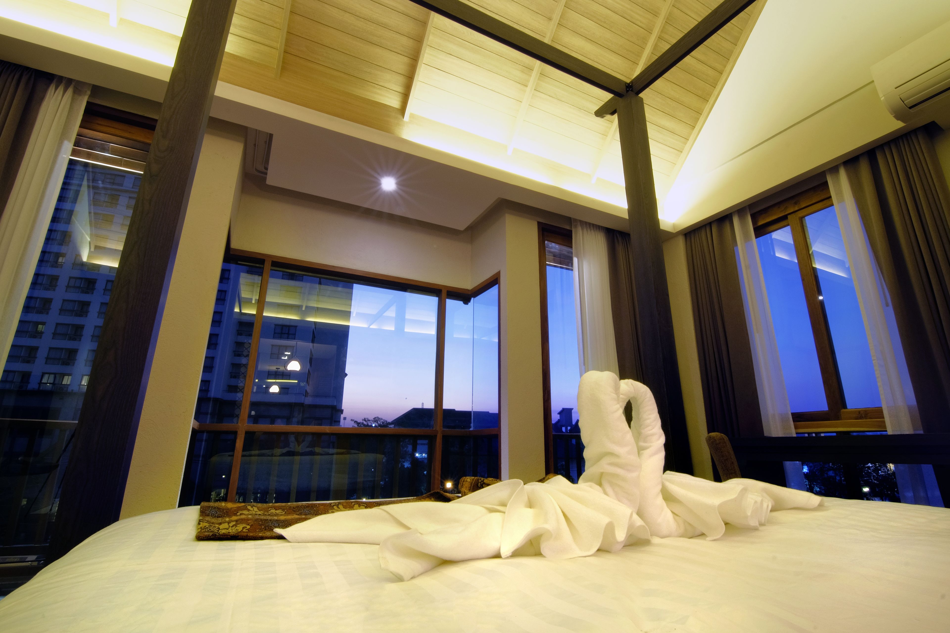 A hotel room with a large bed, a skylight, and a view of a cityscape at dusk.