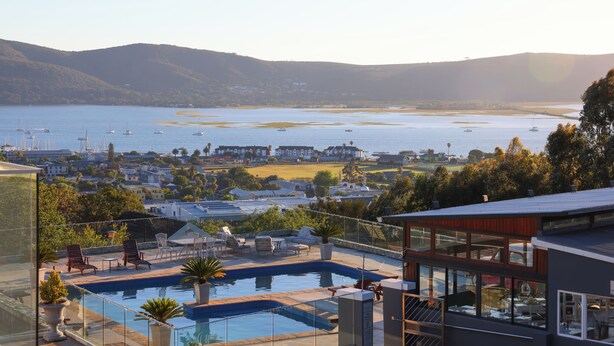 Uma casa moderna com piscina no terraço, com vista para uma baía com barcos e colinas.