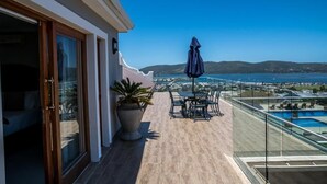 Uma varanda com guarda-corpo de vidro, espaço de estar com mesa e cadeiras, e vista para o mar e colinas.