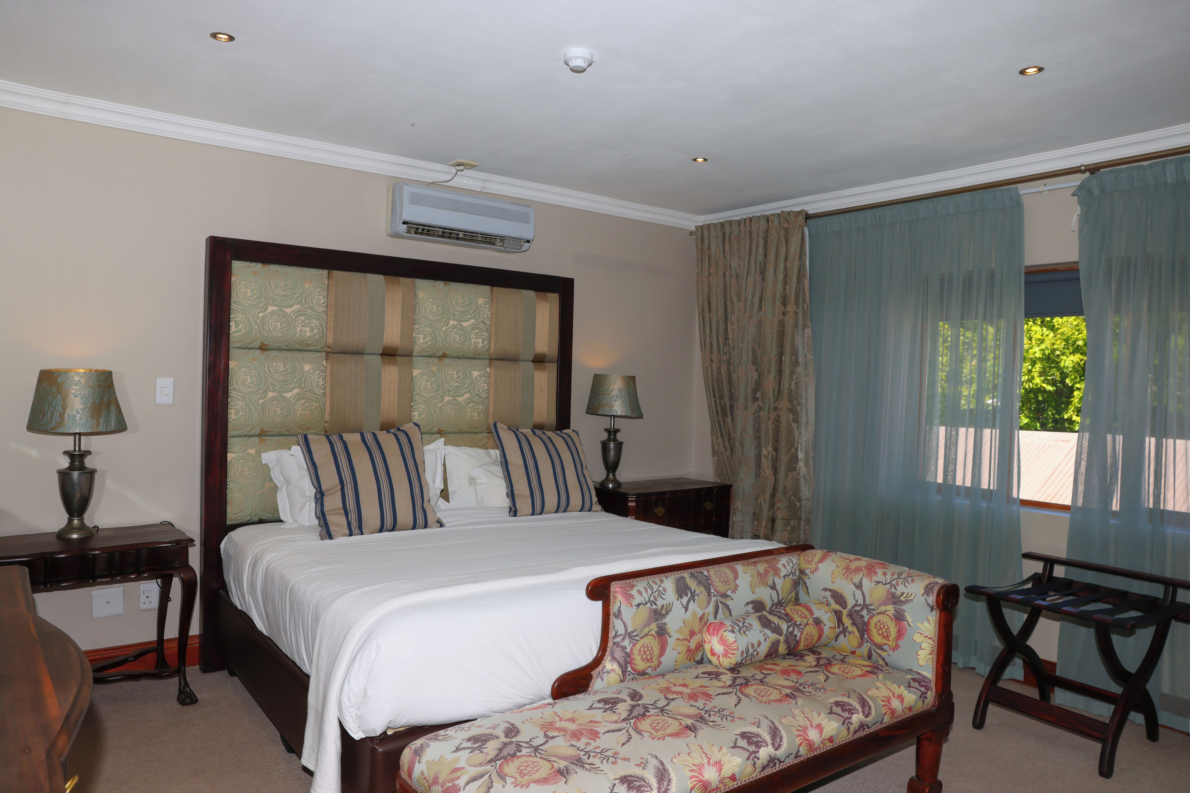 A bedroom with a large bed, two bedside tables with lamps, a floral sofa, and a window with curtains.