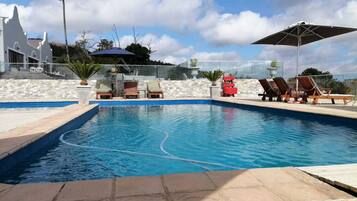 A swimming pool with lounge chairs and a covered area, surrounded by a stone wall and a white building.