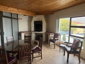 A dining area with a round table and wooden chairs, a fireplace, and a large window with a view of buildings and trees. - Hermanus Heights Villa (Hermanus)
