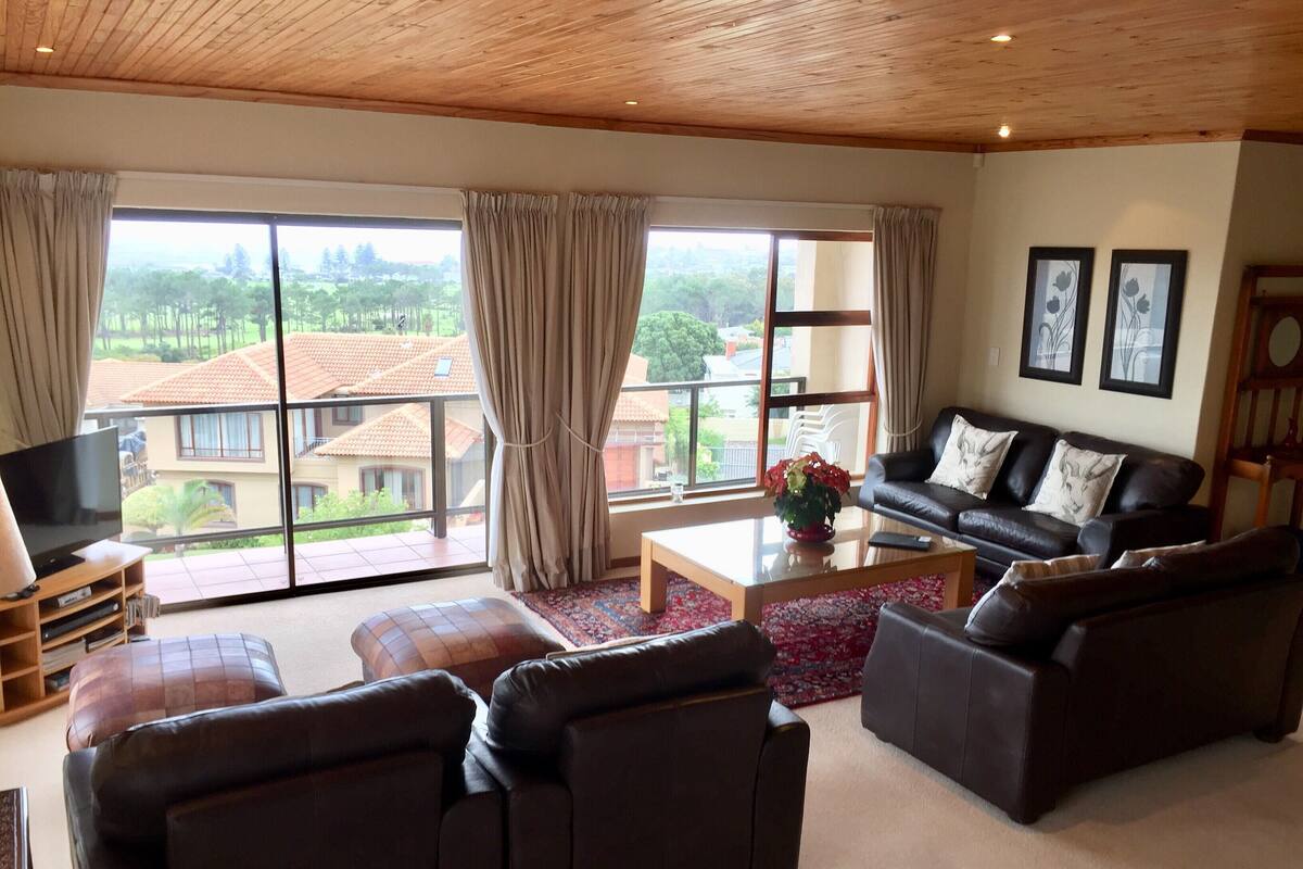 A spacious living room with a large window, a flat-screen TV, and a black leather sofa set.