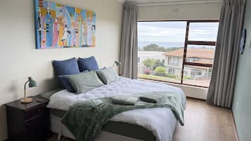 Un dormitorio con una cama grande, una mesita de noche, una lámpara, un cuadro y vistas al exterior.