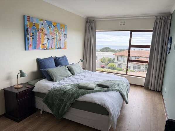 Un dormitorio con una cama grande, una mesita de noche, una lámpara, un cuadro y vistas al exterior.