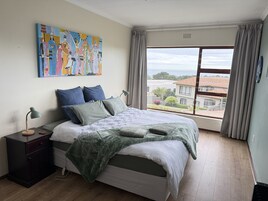 Un dormitorio con una cama grande, una mesita de noche, una lámpara, un cuadro y vistas al exterior.