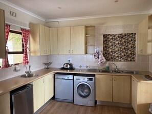 A compact kitchen with wooden cabinets, a Bosch washing machine, a sink, and a window with red curtains. - Hermanus Heights Villa (Hermanus)