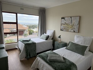 A hotel room with two beds, a nightstand, a window with a view of buildings, and a painting on the wall. - Hermanus Heights Villa (Hermanus)