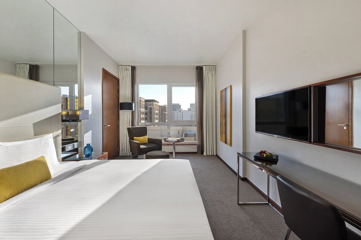 A hotel room with a large bed, a desk with a flat-screen TV, a chair, and a view of the city through large windows.