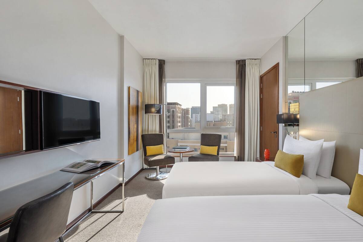 A hotel room with a large bed, a desk with a flat-screen TV, two armchairs, a small table, and a view of the city through large windows.