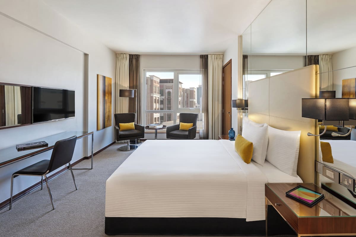A hotel room with a large bed, two armchairs, a desk with a flat-screen TV, and a large window offering a city view.