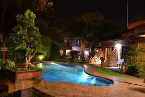 Un espace piscine nocturne avec une terrasse en briques, entouré d’arbres et d’un bâtiment doté d’un éclairage extérieur.