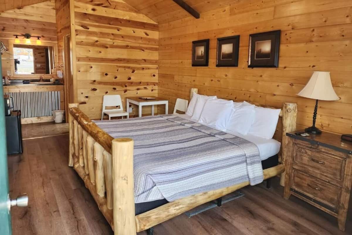 Quarto de cabana de madeira com uma cama, uma pequena mesa, uma cadeira, um abajur e um armário de madeira.