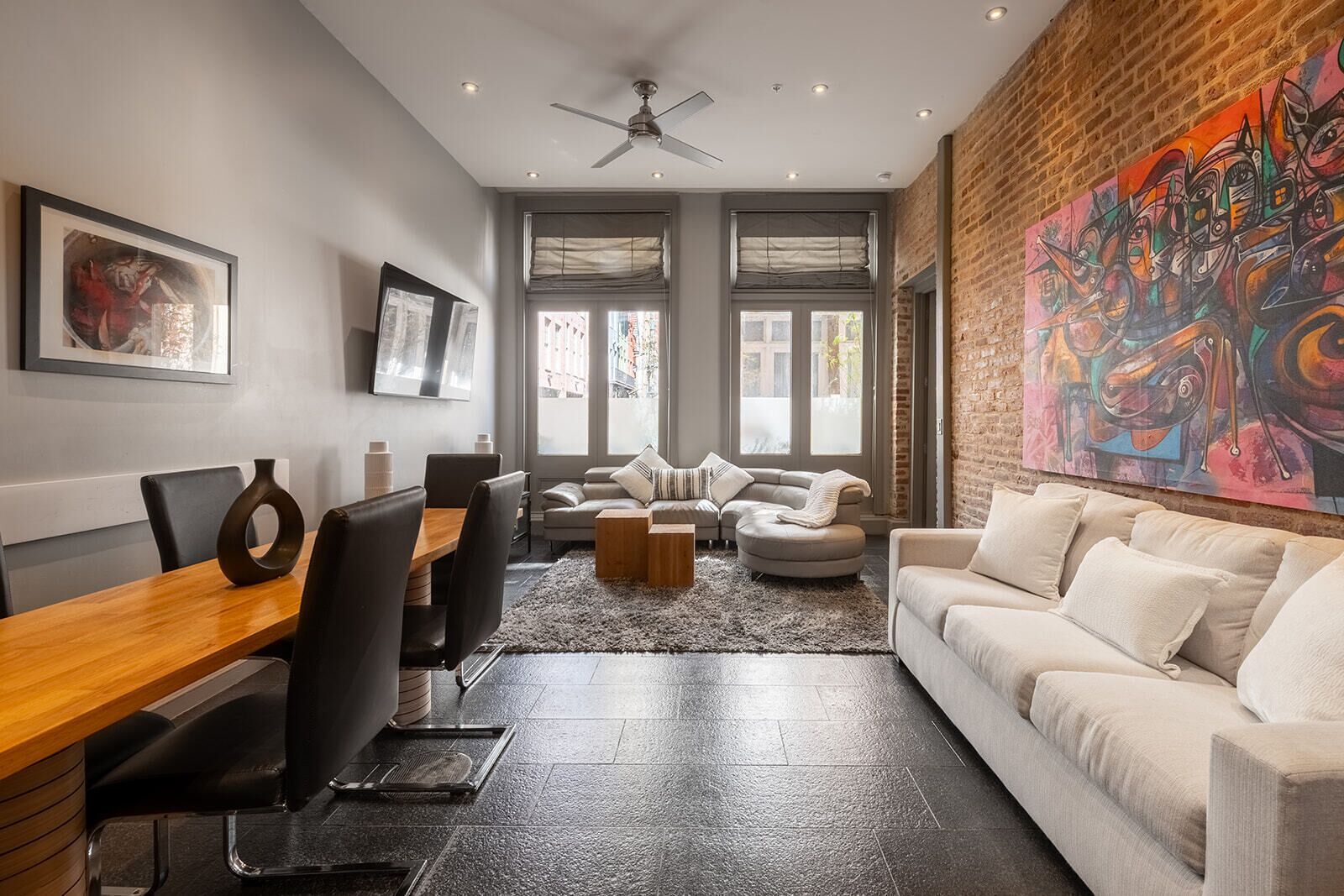 A modern living room with a dining area, featuring a brick wall, large windows, and a ceiling fan.
