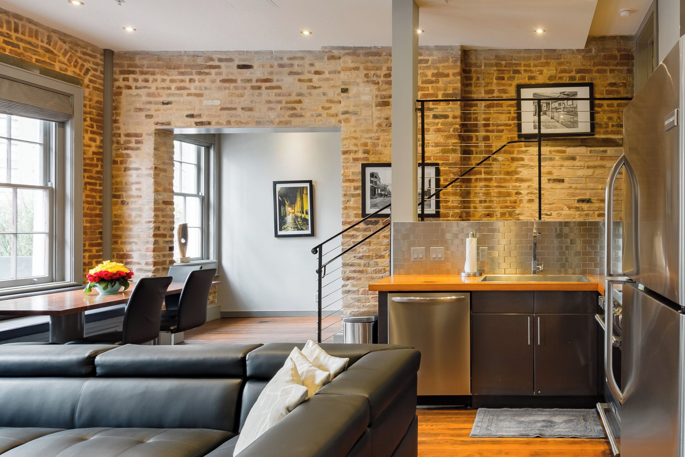 A modern kitchen with a brick wall, stainless steel appliances, and a dining area with a leather sofa and a vase of flowers.