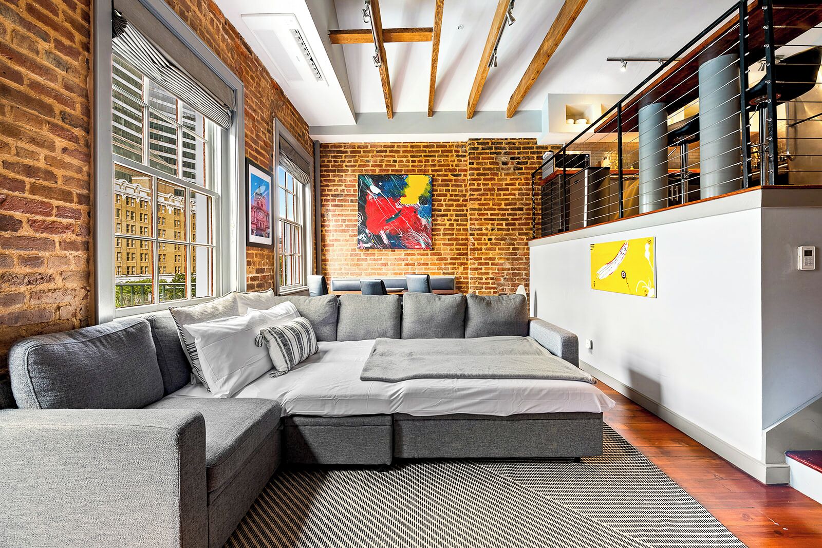 A modern living room with a sectional sofa, exposed brick walls, and a staircase with a metal railing.