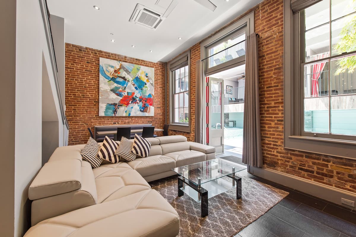 A modern living room with a sectional sofa, a glass coffee table, and a large abstract painting on the brick wall.