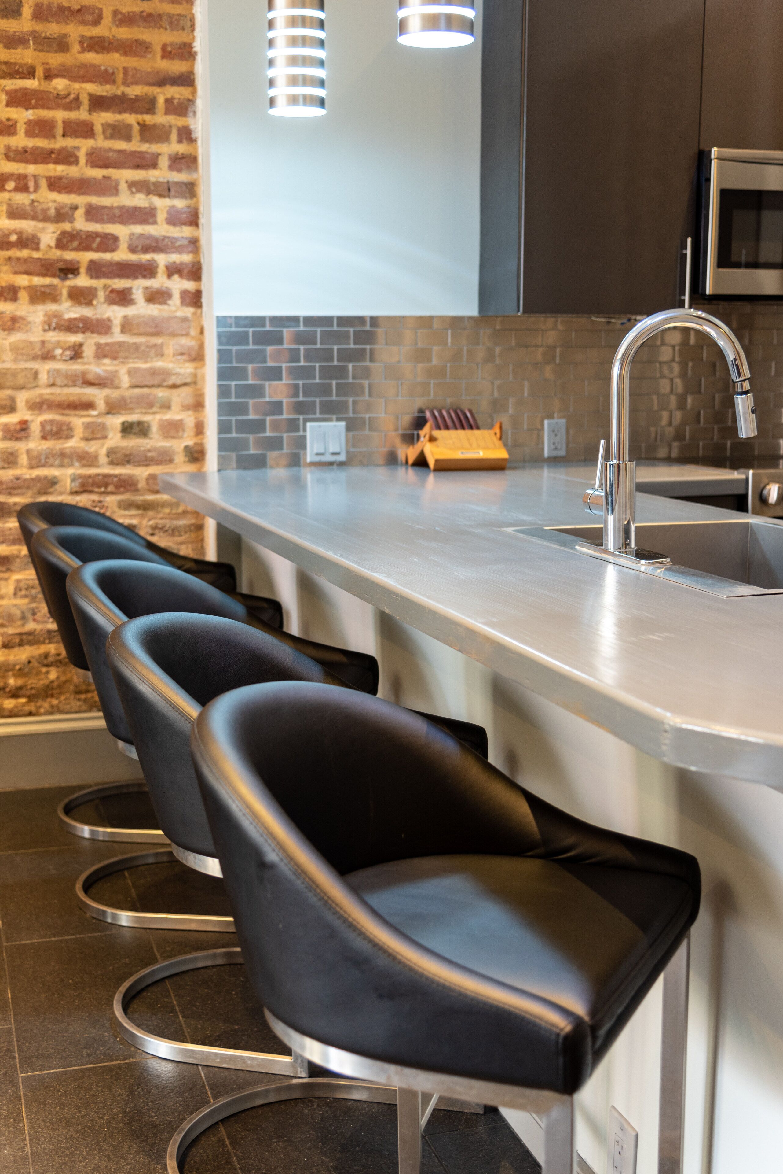 A modern kitchen with a brick wall, a stainless steel bar with four black barstools, a built-in sink, and a microwave.