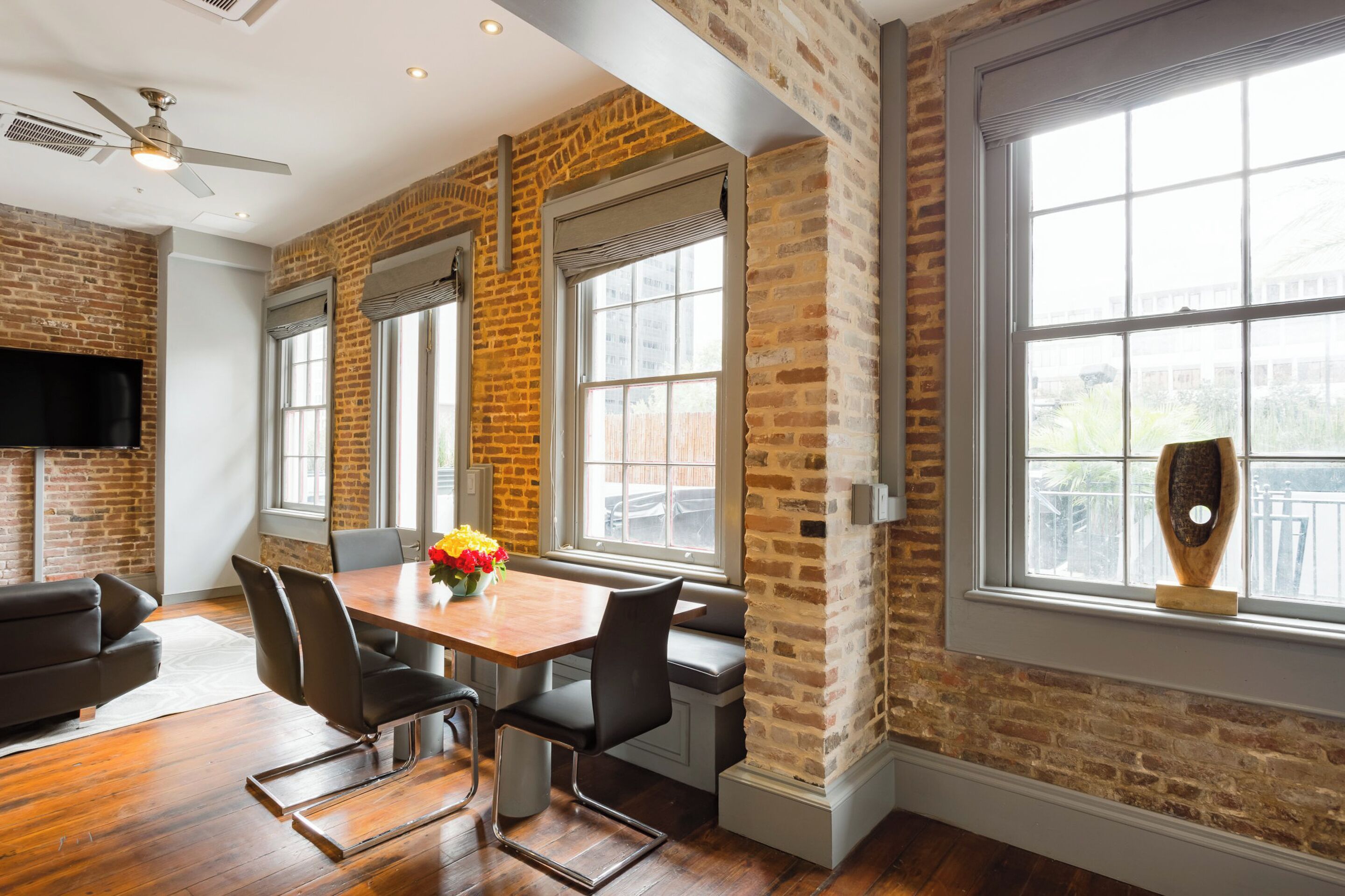 A dining area with a brick wall, large windows, a ceiling fan, and a flat-screen TV.