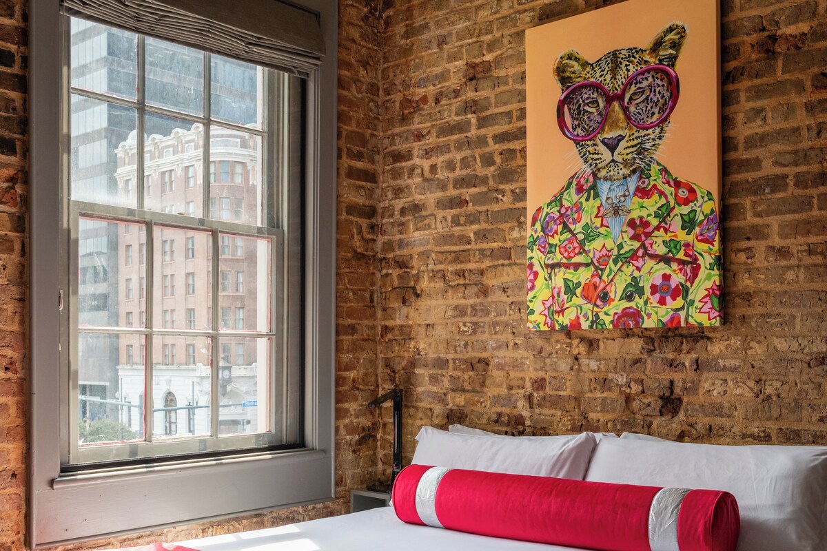 Une chambre d’hôtel avec un mur en briques, une grande fenêtre, un lit avec une literie blanche et rouge, et un tableau représentant un léopard accroché au mur.