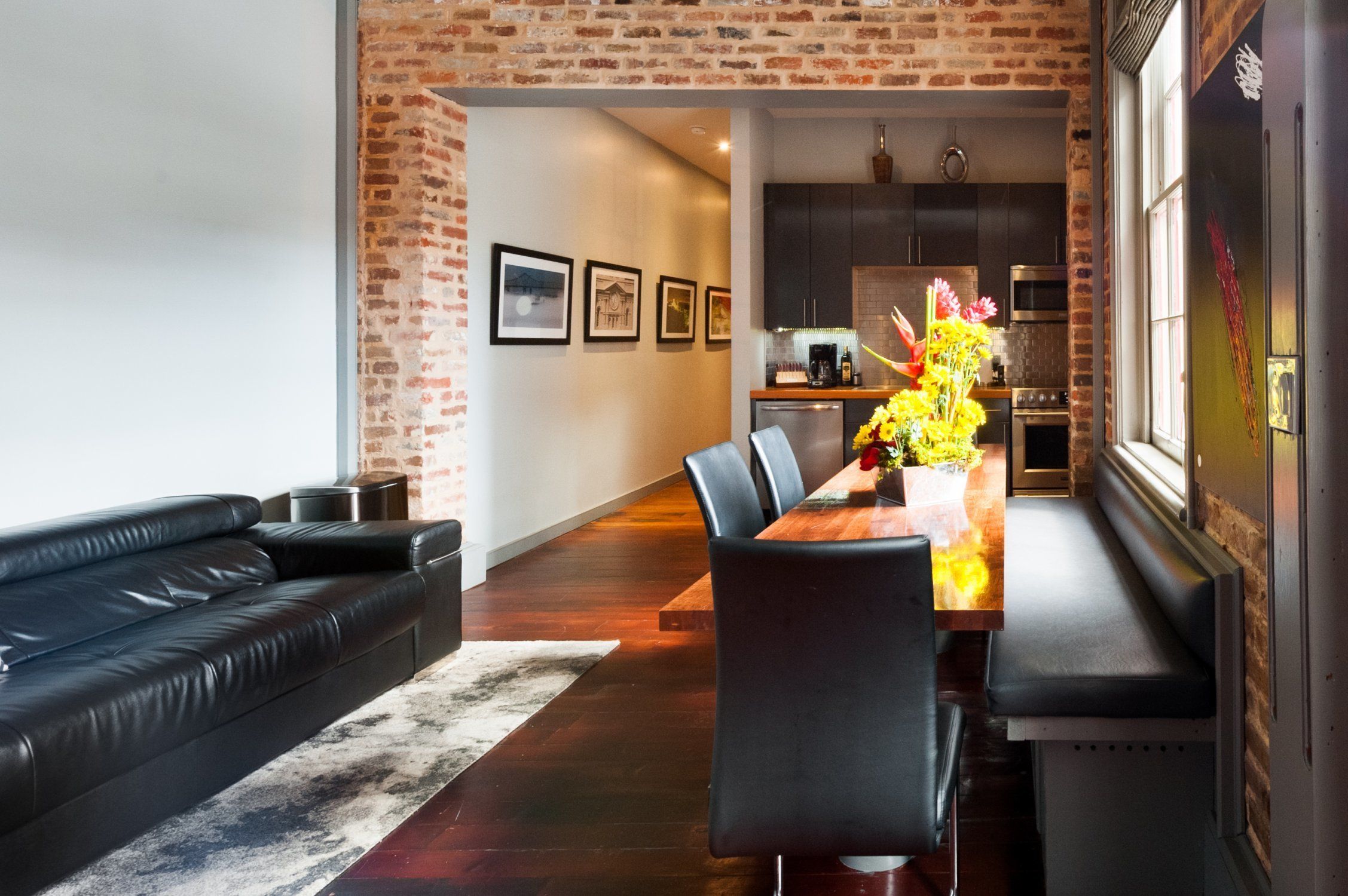 Un salon moderne avec un canapé en cuir noir, une table à manger en bois et des chaises, et un mur en briques.
