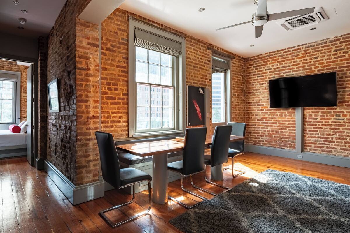 A modern room with exposed brick walls, a wooden floor, a large window, a flat-screen TV, and a dining area with chairs.