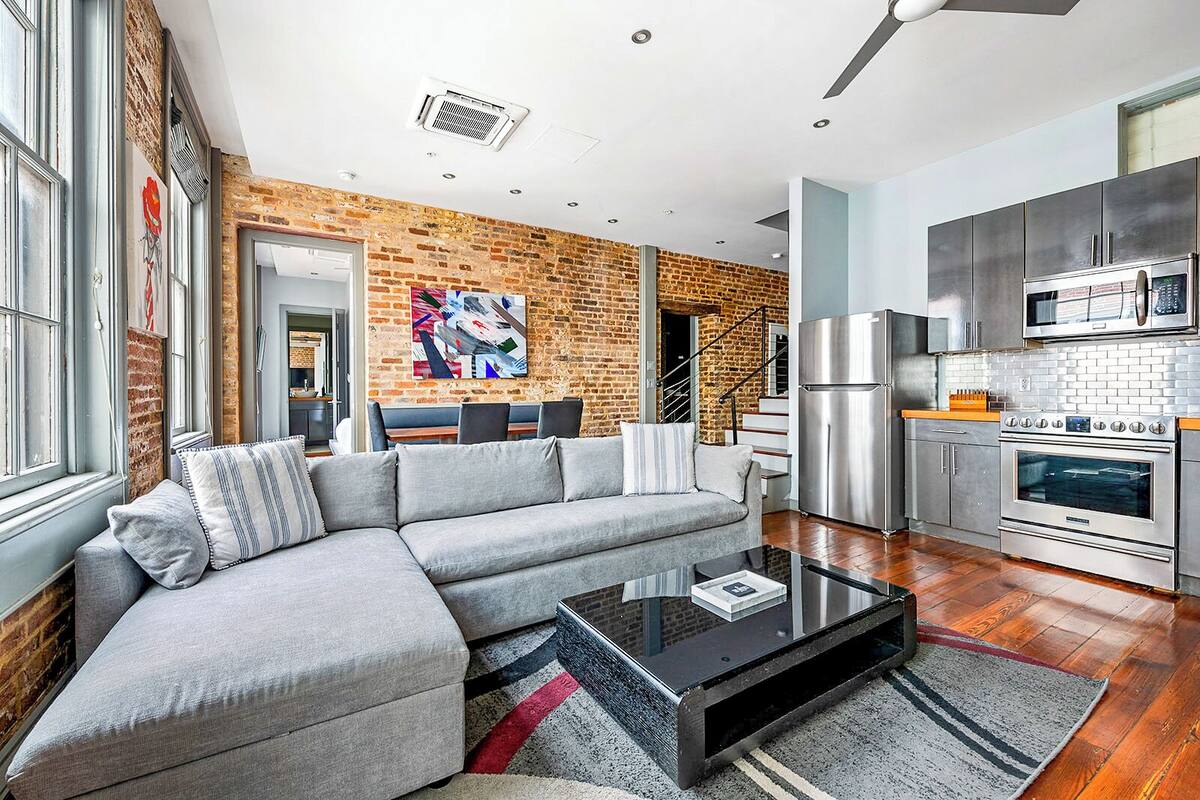 A modern kitchen with stainless steel appliances, a gray sectional sofa, and a brick accent wall.