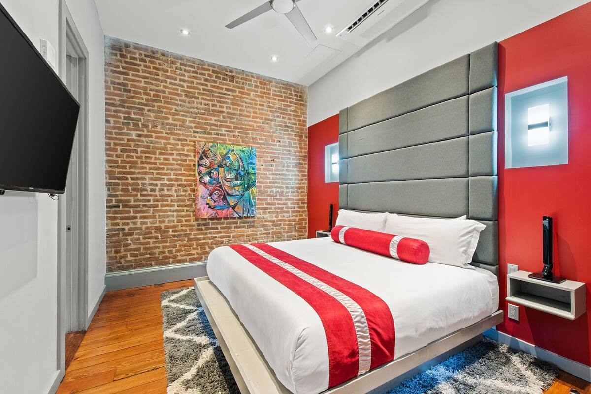 A modern bedroom with a brick wall, a large bed with red and white bedding, a grey headboard, and a ceiling fan.
