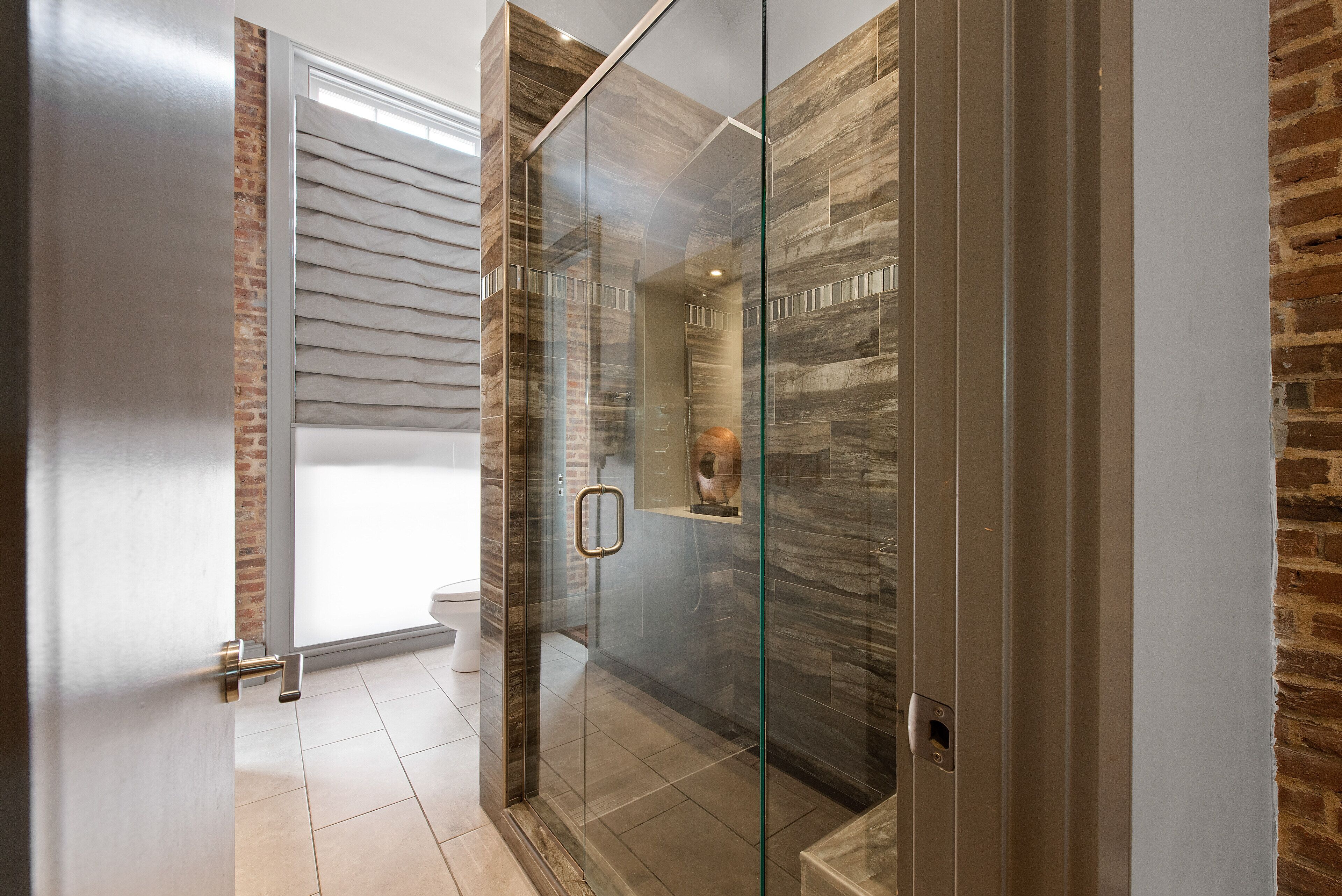 Un baño moderno con ducha de vidrio, inodoro a la vista y pared decorativa de ladrillo.