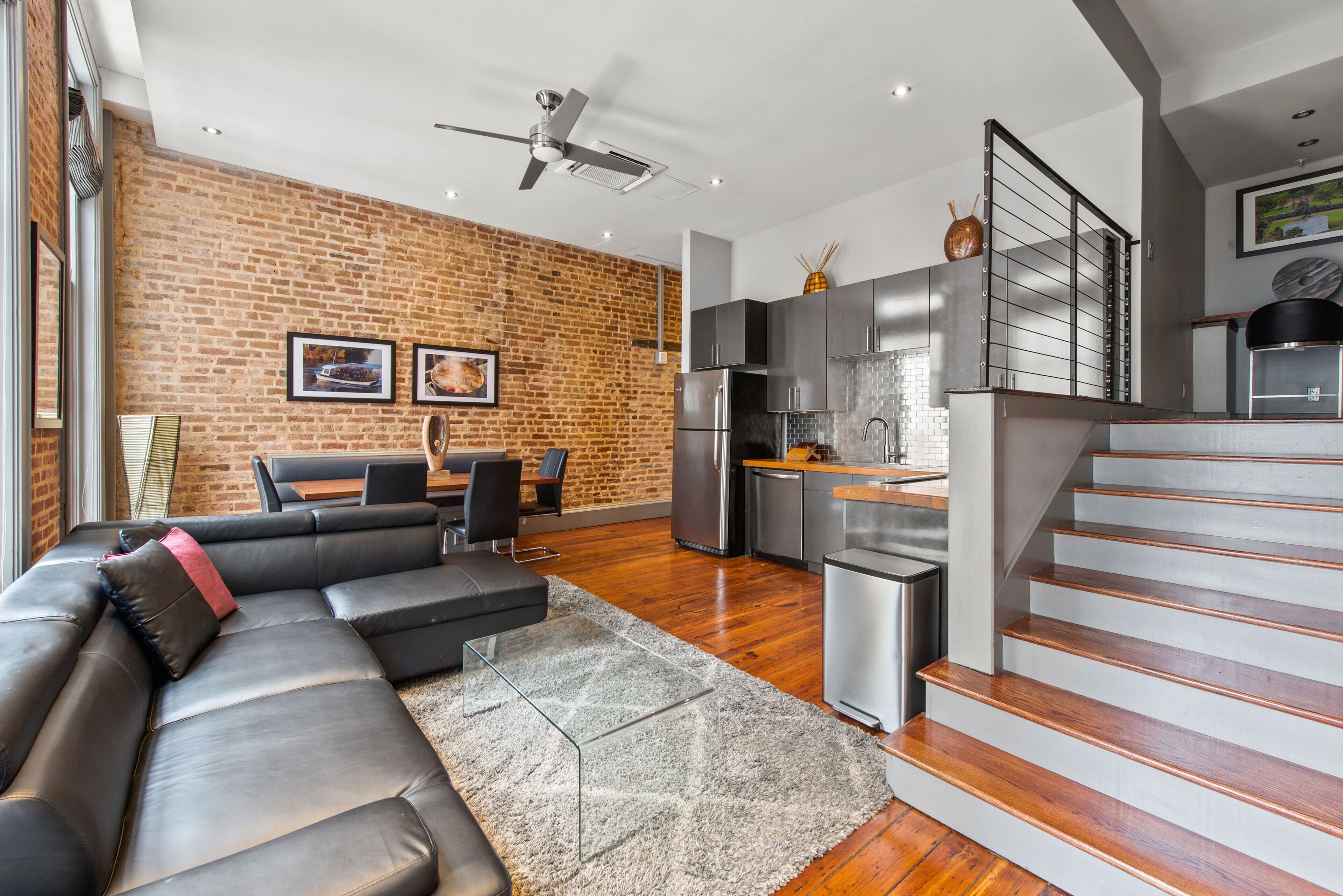 A modern living room with a sectional sofa, a dining area, a kitchen with stainless steel appliances, and a staircase.
