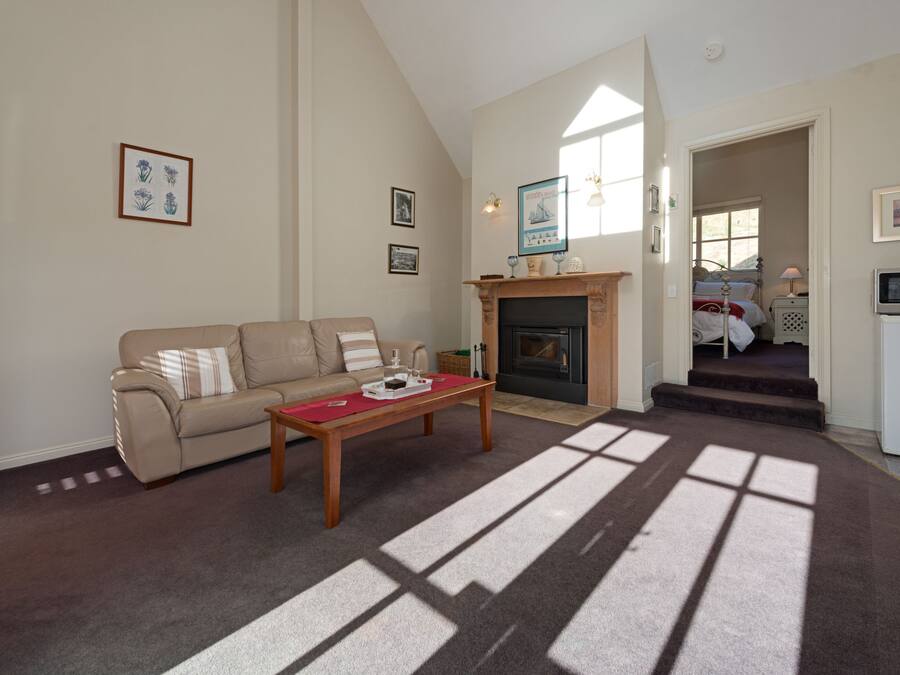 A living room with a sofa, a coffee table, and a fireplace. There are framed pictures on the wall and a window allowing natural light.