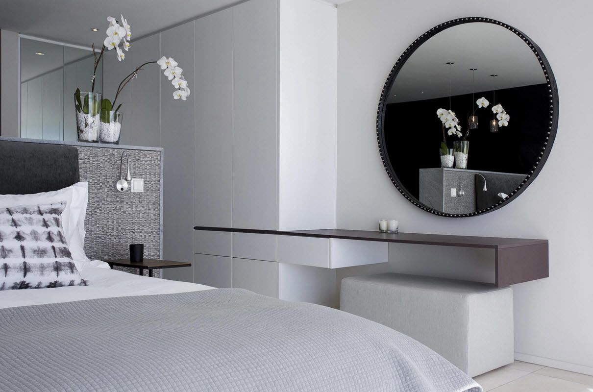 A modern bedroom with a large bed, a bedside table, a vanity with a mirror, and orchid plants.
