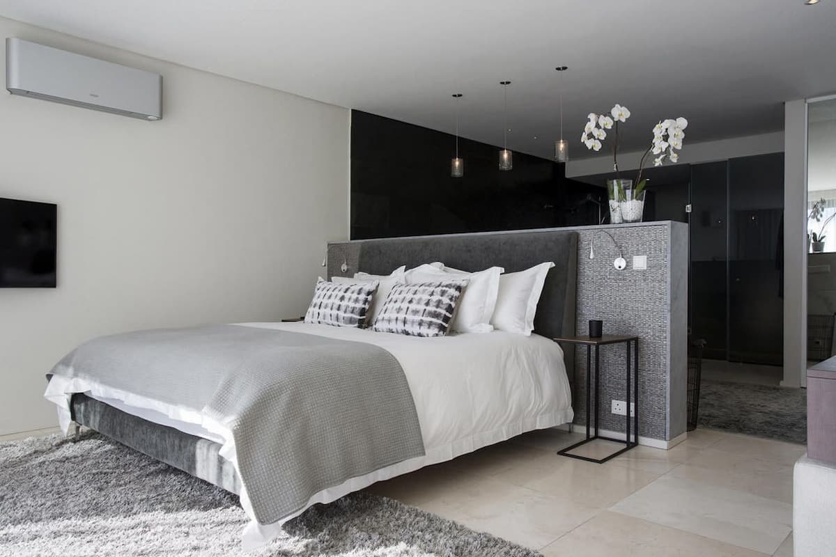Un dormitori modern amb un llit gran, una combinació de colors grisos i blancs, una petita taula auxiliar i un aire condicionat fixat a la paret.