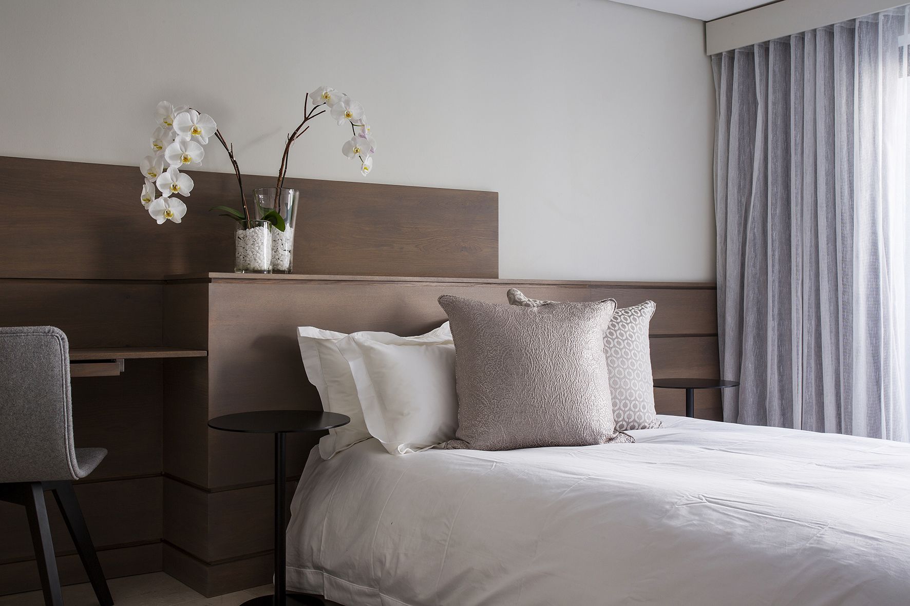 Hotelkamer met een bed, twee kussens, een nachtkastje, een stoel en een vaas met witte bloemen.