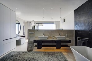 A modern bathroom with a large mirror, two sinks, a bathtub, and a large window.