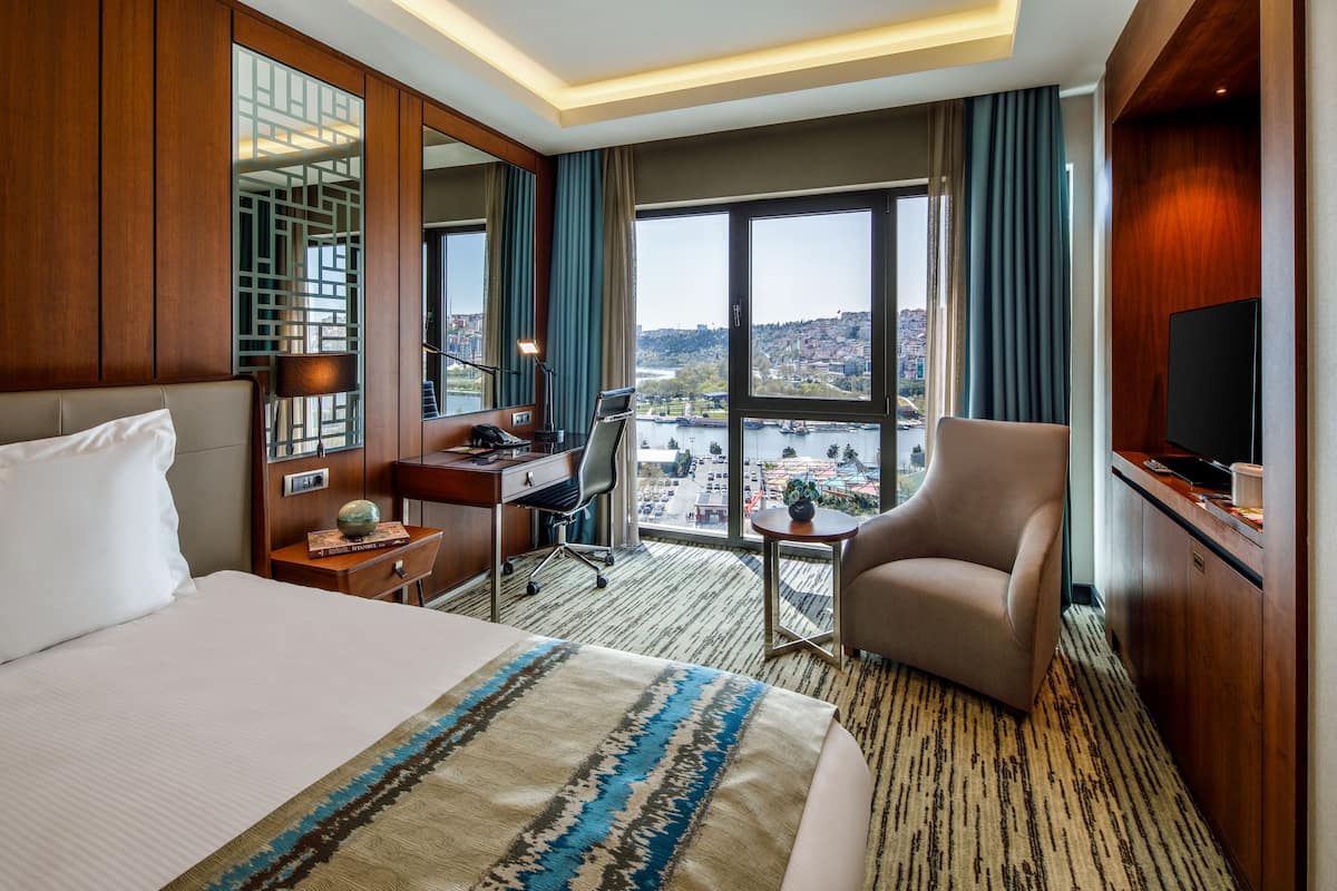 A hotel room with a large bed, a desk, a chair, a TV, and a view of a cityscape through large windows.