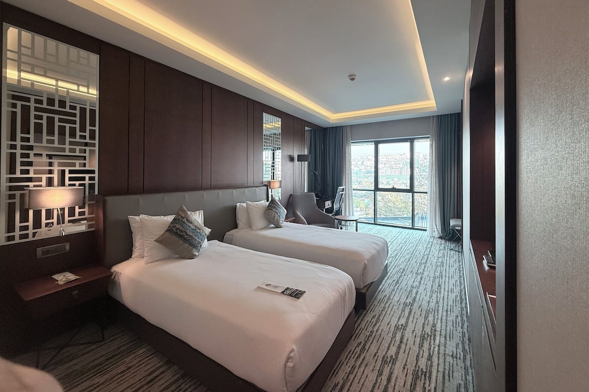 A hotel room with two beds, a desk, a chair, a lamp, and a view of the city through large windows.