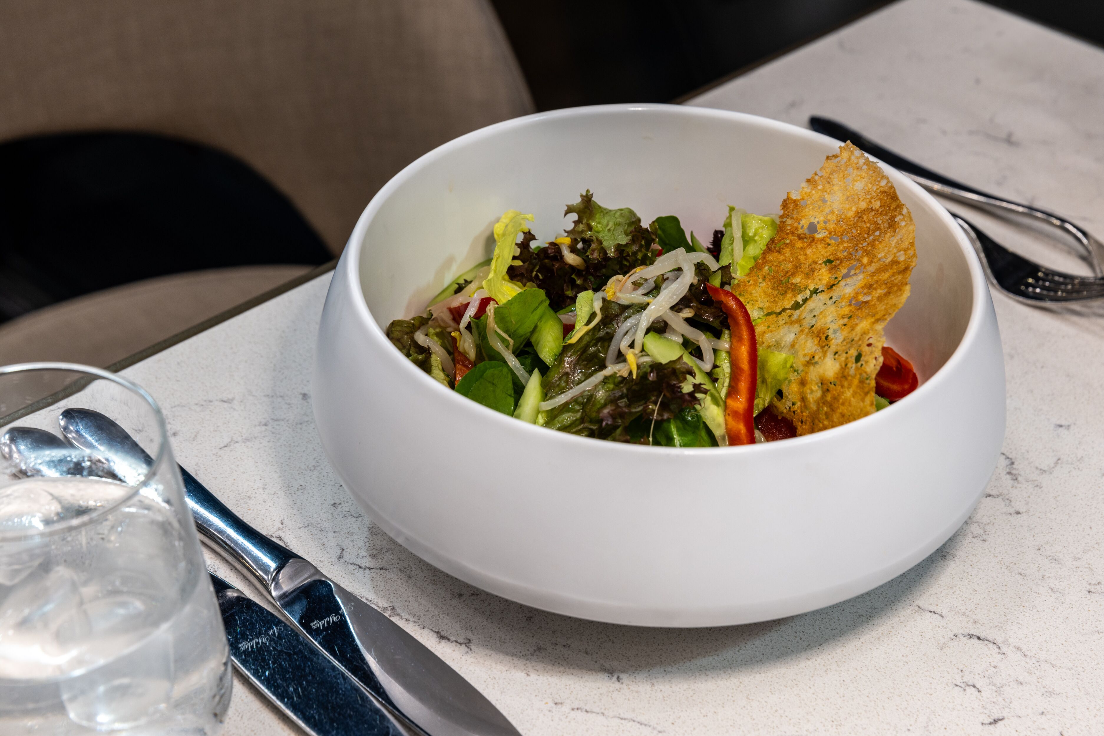 A white bowl of salad with a crispy bread piece on a marble table.