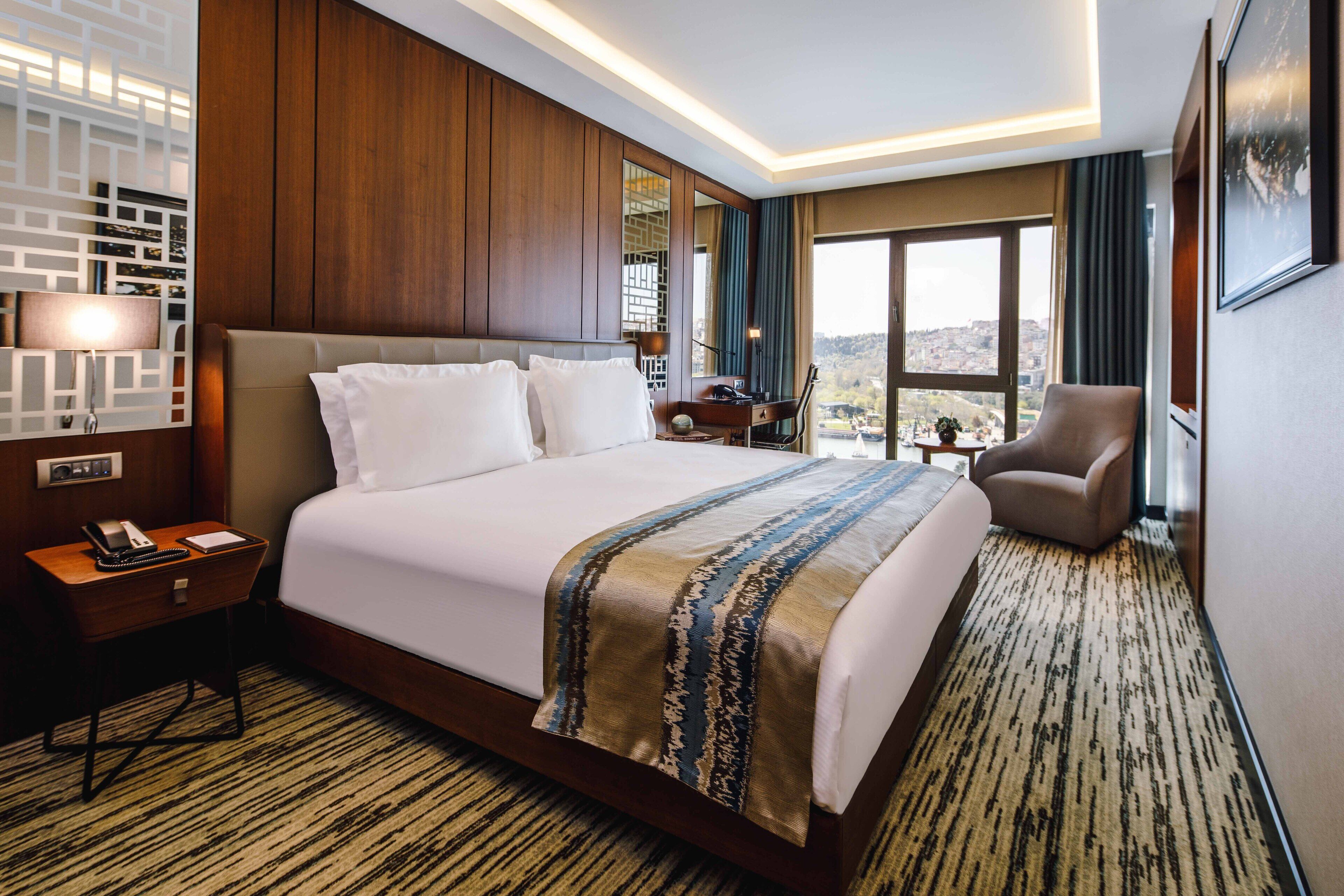 A hotel room with a large bed, a desk with a lamp, a chair, and a view of the outdoors.